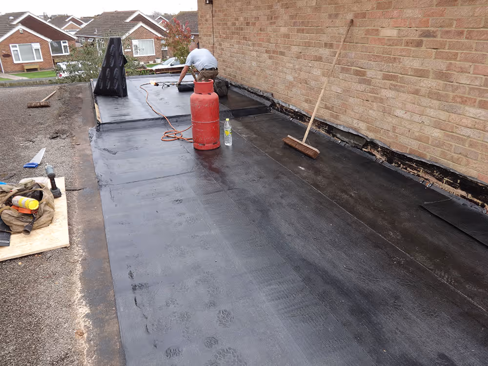 Worker applying roofing material on flat roof next to wall, with a broom and gas cylinder nearby.